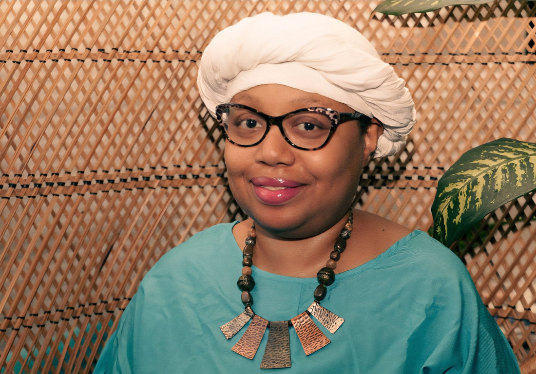 Idi E. Utuk, founder of Regulate Wellness, wearing a white headwrap and teal top against a wicker background.
